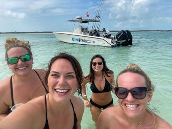 Four smiling women in bikinis taking a selfie while standing in clear turquoise shallow water at a tropical sandbar, with a white motorboat anchored nearby under a partly cloudy sky.