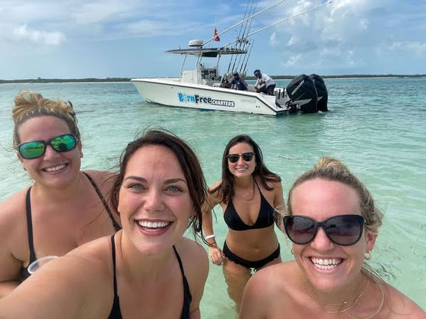 Four smiling women in bikinis taking a selfie while standing in clear turquoise shallow water at a tropical sandbar, with a white motorboat anchored nearby under a partly cloudy sky.