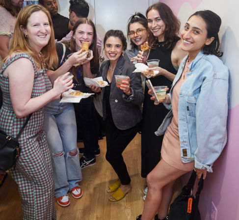 Six friends in casual outfits laughing and eating pastries while holding drinks at a cozy indoor gathering by a pink wall.