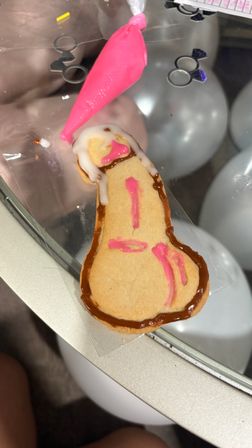 Novelty penis-shaped sugar cookie on a glass table being decorated with white and hot-pink icing from a piping bag, brown icing border and blurred balloons in the background