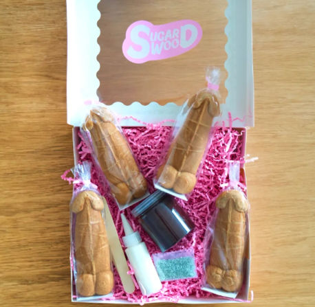 Adult bachelorette cookie decorating kit on wooden table: white gift box with pink crinkle paper holding four individually wrapped penis-shaped cookies, small jar of chocolate, icing bottle, sprinkles packet and wooden spatula.