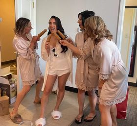 Bridal party getting ready in a hotel suite — laughing bride in white robe and fluffy slippers surrounded by three bridesmaids in matching blush robes playfully offering corn dogs.