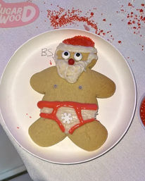 Holiday gingerbread cookie decorated as a cheeky Santa with white icing beard, red sprinkle hat and red-iced underwear on a white plate with scattered red sprinkles