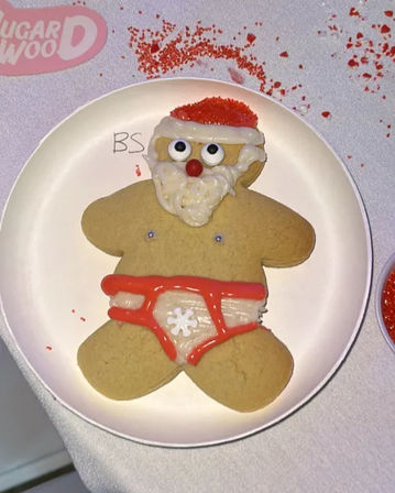 Holiday gingerbread cookie decorated as a cheeky Santa with white icing beard, red sprinkle hat and red-iced underwear on a white plate with scattered red sprinkles