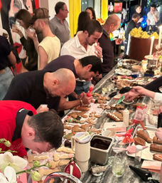 Adults crowded around a marble countertop at an indoor holiday party, busy decorating gingerbread cookies with icing, piping bags, toppings and drinks nearby.