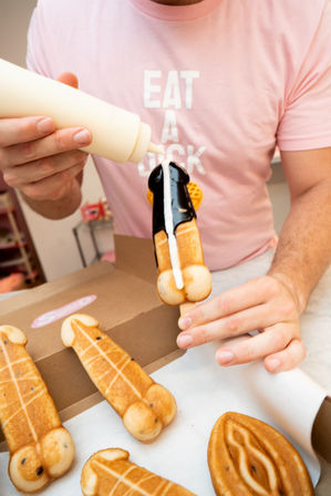 Hands holding a novelty penis-shaped waffle on a stick being drizzled with chocolate and white sauce, with a person in a pink T‑shirt in the background and a box of similar cheeky-shaped waffles on the counter.