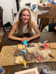 Smiling bride-to-be wearing a pearl "BRIDE" headband and veil at a wooden kitchen table with cookie decorating supplies — icing bags, sprinkles and shaped cookies for a bridal shower
