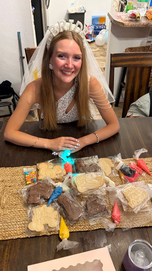 Smiling bride-to-be wearing a pearl "BRIDE" headband and veil at a wooden kitchen table with cookie decorating supplies — icing bags, sprinkles and shaped cookies for a bridal shower