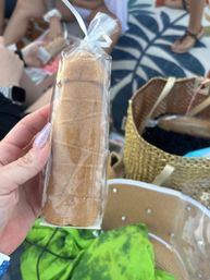 Hand with sparkly purple nails holding a clear-bagged cinnamon-sugar sponge cake roll at a beach picnic, woven tote and tropical leaf towel in the background.