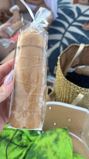 Hand with sparkly purple nails holding a clear-bagged cinnamon-sugar sponge cake roll at a beach picnic, woven tote and tropical leaf towel in the background.
