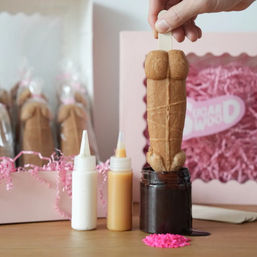 Playful adult-themed novelty cookie pop on a stick being dipped in chocolate, with white and caramel icing bottles, pink sprinkles and a pink gift box in the background