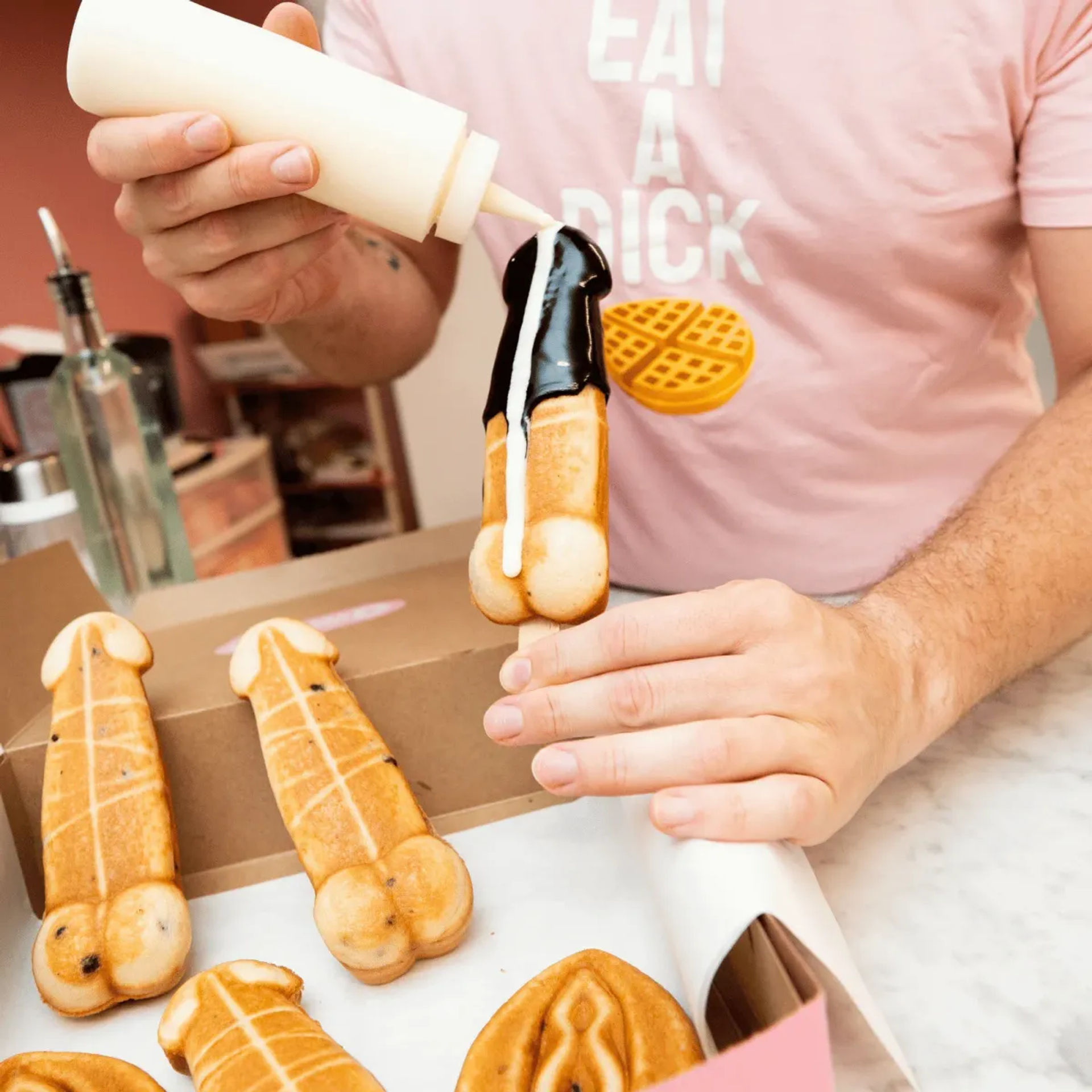 Close-up of hands drizzling chocolate and white sauce over a novelty phallic-shaped waffle pop on a stick, with more waffle pops on a bakery counter