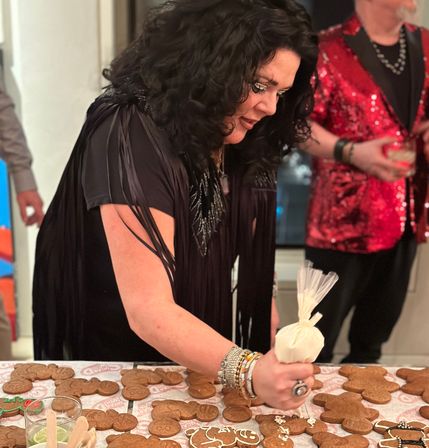 Person in a black fringe top piping icing onto gingerbread cookies at a festive indoor holiday cookie-decorating party, guest in a red sequined jacket visible in the background.