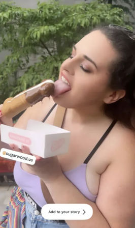 Person in a lavender tank top licking a chocolate-dipped popsicle outdoors, holding a white takeout box on a sunny urban patio — summer street-food treat.