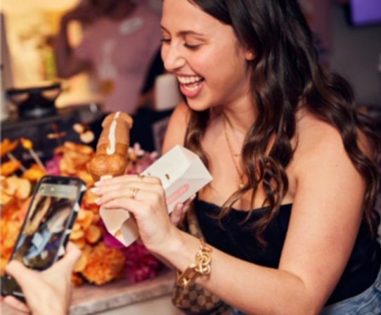 Laughing woman at a dessert tasting holding a cream-filled pastry pop while a friend snaps a photo, bright floral display behind
