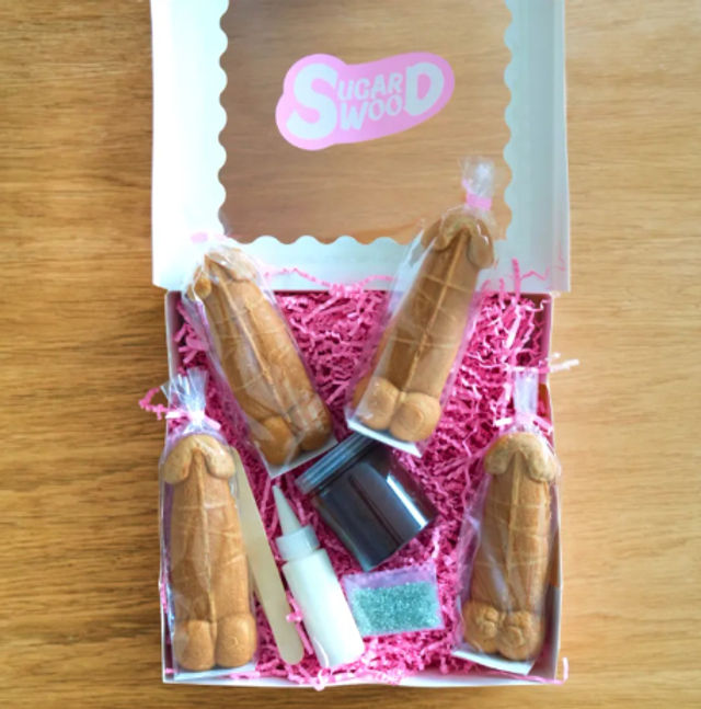 Adult novelty cookie decorating kit in a pink shredded-paper gift box on a wooden table, four individually wrapped penis-shaped molded cookies with a small frosting bottle, chocolate jar, sprinkles packet and wooden spatula.