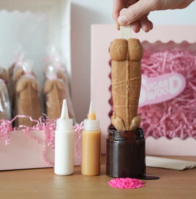 Hand holding a novelty phallic waffle pop on a wooden stick being dipped into melted chocolate, with white and caramel sauce bottles, pink gift box and pink sprinkles on a table.