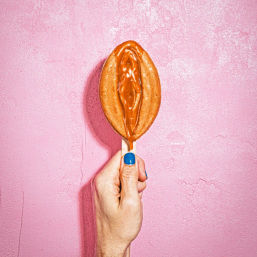 Close-up of a hand with blue nail polish holding a caramel-filled oval waffle pop on a stick, caramel dripping, set against a pink textured wall