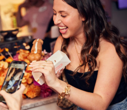 Smiling woman at an indoor party holding a chocolate éclair in a takeaway box while someone photographs it with a smartphone at a colorful dessert table.