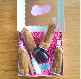 White gift box on wooden table filled with pink paper shred holding five individually wrapped novelty penis-shaped cookies and a small decorating kit (icing bottle, sprinkles packet, wooden stick) — playful bachelorette party treat.