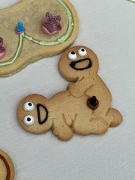Close-up of decorated gingerbread cookies on a light linen surface: a playful double gingerbread figure with candy eyes and black-icing smiles, next to part of a star-topped cookie with pastel frosting.