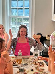 Joyful woman in a pink apron holds a decorated sugar cookie at a sunlit indoor cookie-decorating party, surrounded by smiling guests, iced cookies, bowls, glasses and water bottles on a busy table.