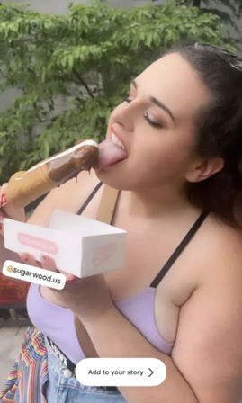 Person outdoors enjoying a chocolate-dipped ice cream bar on a stick, holding a white takeout box, wearing a lilac top and denim shorts against a leafy patio background.