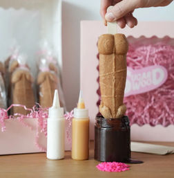 Hand holding a cheeky adult-shaped cookie pop being dipped into a jar of chocolate sauce on a wooden table, with white and caramel squeeze bottles, pink sprinkles, and gift boxes filled with shredded paper in the background.