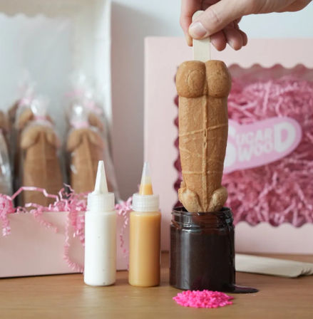 Hand holding a cheeky adult-shaped cookie pop being dipped into a jar of chocolate sauce on a wooden table, with white and caramel squeeze bottles, pink sprinkles, and gift boxes filled with shredded paper in the background.