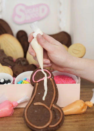 Hand piping white icing onto an adult-themed chocolate cookie with pink and brown icing accents at a cookie-decorating station, surrounded by bowls of sprinkles and colorful piping bags on a wooden table