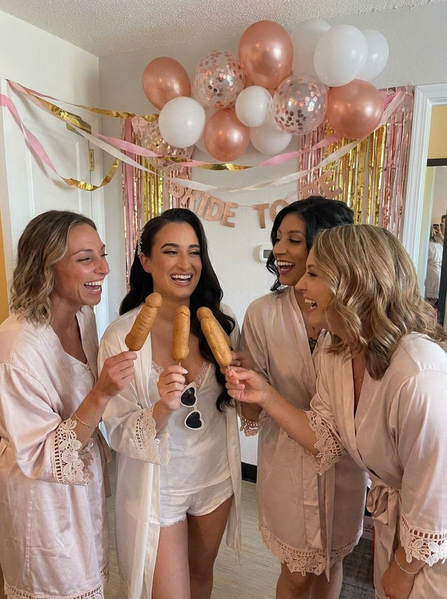 Four women in blush satin robes laughing and toasting corn dogs at an indoor rose-gold themed bridal shower with balloons and a "Bride to Be" banner