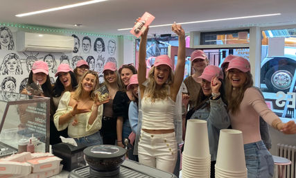 Cheerful group of women in matching pink caps celebrating inside a bright dessert shop, holding decorated cookies and snapping photos by a muraled wall and counter with coffee cups and pastry boxes.