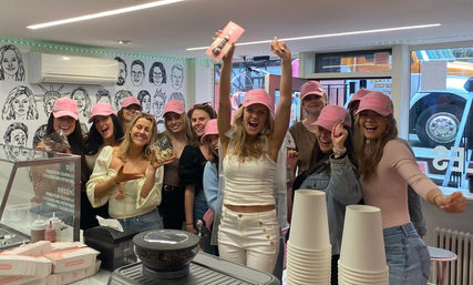 Group of smiling women wearing matching pink baseball caps celebrating at a bright dessert shop counter, cheering and holding decorated cookies amid pastry boxes, stacked takeaway cups, and illustrated wall art.