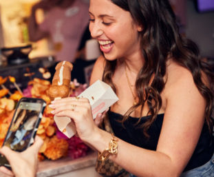 Smiling woman at a dessert stand holding a chocolate-drizzled pastry on a stick while a friend photographs it with a smartphone, colorful flowers blurred in the background