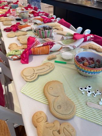 Kids' cookie-decorating party setup in a home kitchen: long table lined with plain shaped sugar cookies, bowls of gumdrops and candy eyes, small sugar bowls, and colorful napkins tied with bright pink bows.