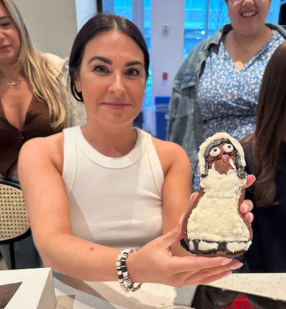 Smiling woman in a white tank top holding a whimsical cream-covered pastry shaped like a doll, friends gathered around a bright indoor café or bakery counter.