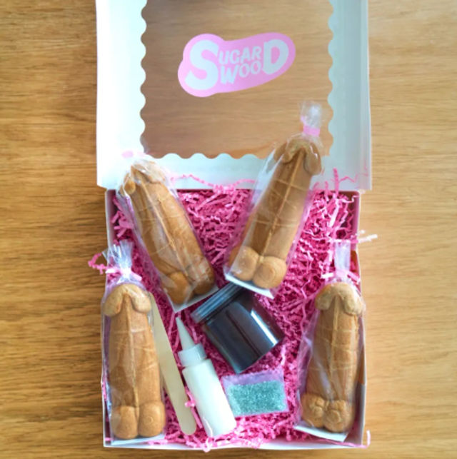Open gift box on wooden table containing a novelty cookie-decorating kit: four penis-shaped cookies individually wrapped on pink crinkle paper with a small icing bottle, chocolate jar and sprinkles.