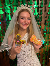 Smiling bride-to-be wearing a pearl "BRIDE" headband and veil, sequined dress, holding two colorful novelty adult-themed cookies against a backdrop of green hanging decorations at a bachelorette-style celebration.