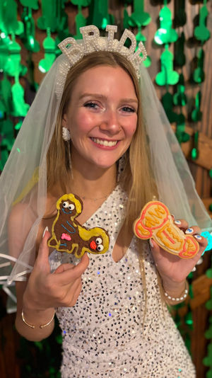 Smiling bride-to-be wearing a pearl "BRIDE" headband and veil, sequined dress, holding two colorful novelty adult-themed cookies against a backdrop of green hanging decorations at a bachelorette-style celebration.