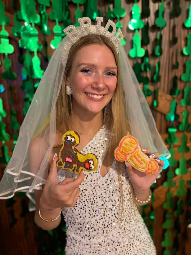 Smiling bride-to-be wearing a pearl "BRIDE" headband and veil, sequined dress, holding two colorful novelty adult-themed cookies against a backdrop of green hanging decorations at a bachelorette-style celebration.