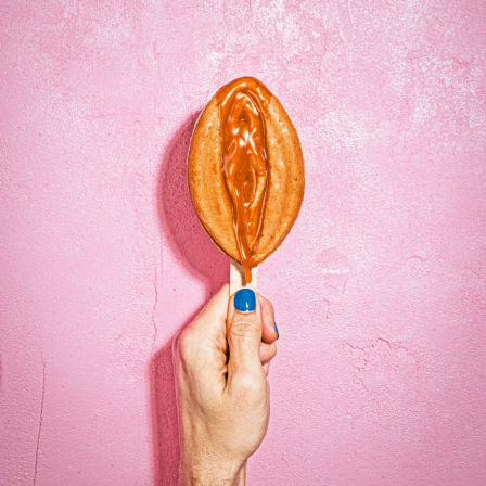 Hand with blue nail polish holding a caramel-drizzled waffle pop on a wooden stick against a textured pink wall.