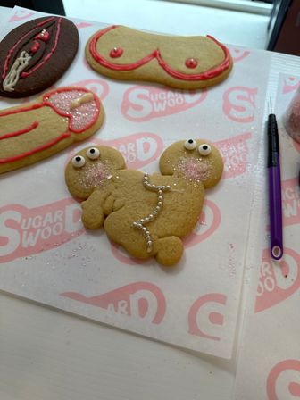 Playful adult novelty sugar cookies decorated as breasts, vulva and penis on pink-logo parchment at a cookie-decorating station, topped with edible pearls, pink sanding sugar and a purple decorating tool.
