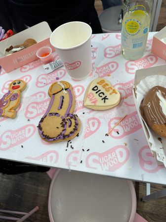 Overhead view of a cookie-decorating table with novelty iced treats — a heart-shaped cookie with an iced message, a phallic-shaped cookie decorated in purple icing and chocolate sprinkles, a paper cup, a clear lemonade bottle, sprinkles and a chocolate-covered pastry on printed paper.