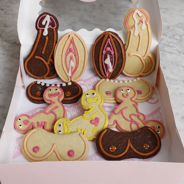 Bakery box of colorful adult-themed novelty sugar cookies — playful penis, vulva, breast and cartoon figures iced in pink, yellow, white and chocolate on a marble countertop.