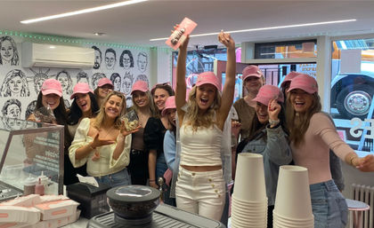 Cheerful group of friends wearing pink caps celebrate a bachelorette at a bright ice cream parlor — bride in white with veil raises her arms over the counter with cups and toppings.