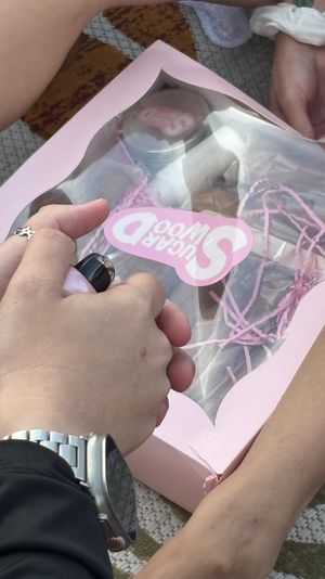 Hands on a patterned blanket opening a pink bakery-style gift box with a clear window and pink crinkle paper; one hand holds a small metallic pink device while legs and a scrunchie-adorned wrist rest nearby.