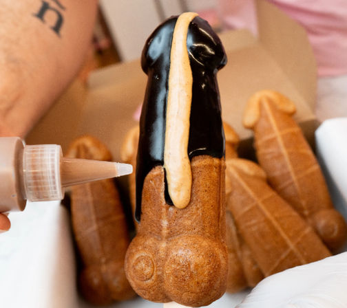 Cheeky close-up of a penis-shaped cream-filled choux pastry topped with dark chocolate glaze and a caramel drizzle — novelty adult bakery dessert.