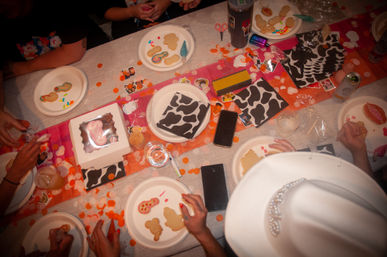Overhead view of a lively cookie-decorating party table: paper plates with plain and iced cookies, piping icing, sprinkles, cow-print napkins, orange confetti, drinks, phones, hands reaching in, and a white rhinestone hat on the edge.