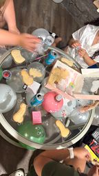 Glass-top party table with silver and green balloons, canned drinks and water, novelty sugar cookies and candy, hands arranging party favors — playful adult party scene.