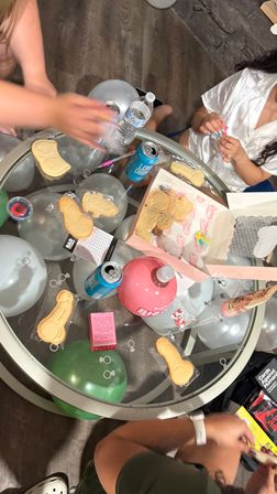 Glass-top party table with silver and green balloons, canned drinks and water, novelty sugar cookies and candy, hands arranging party favors — playful adult party scene.